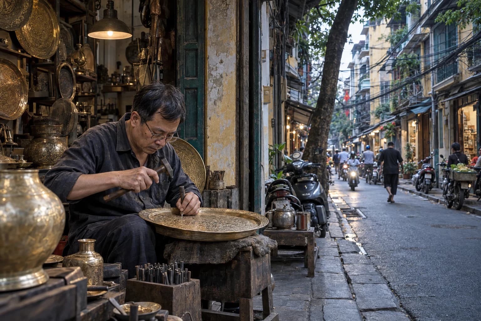 A metal artisan engraving a brass tray in an open-front workshop on a dense Hanoi Old Quarter street